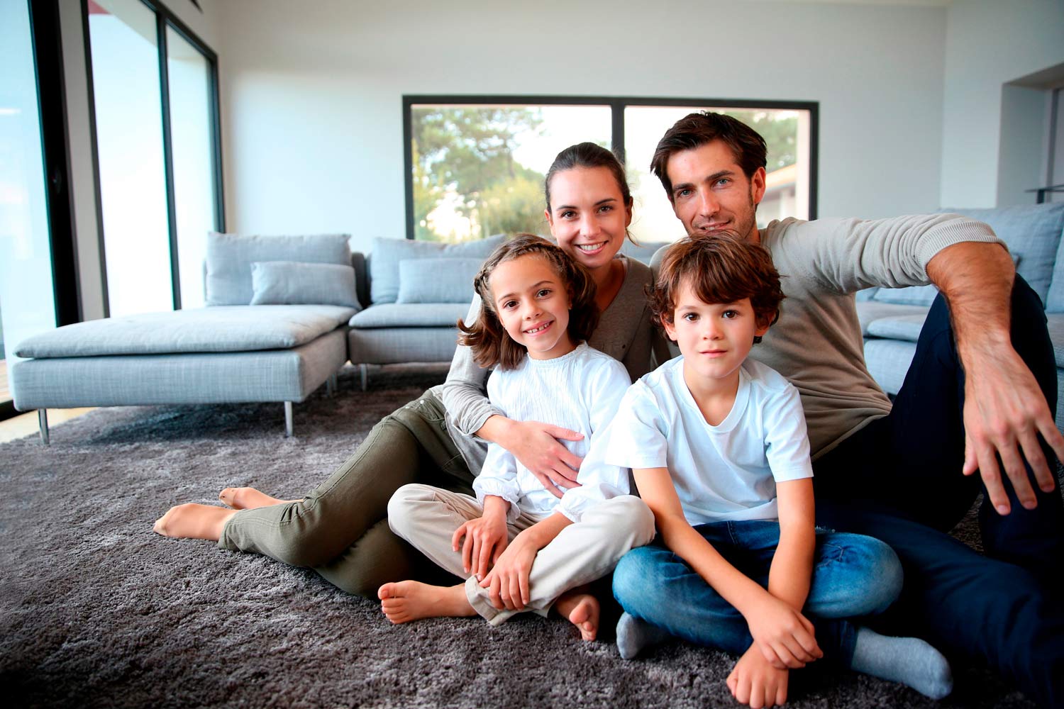Família de pai, mãe, com filho e filha pequenos, sentados no chão da sala à frente de sofá.