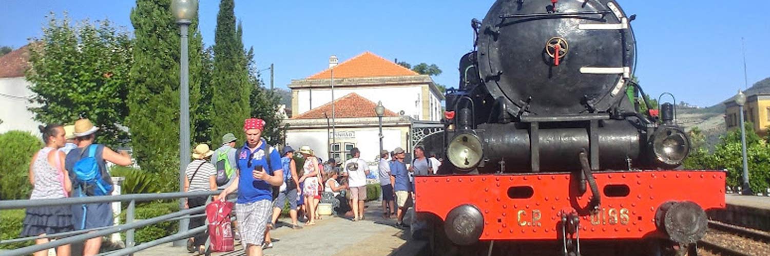 Frente da locomotiva do Comboio Histórico do Douro na estação do Pinhão e passageiros na plataforma.
