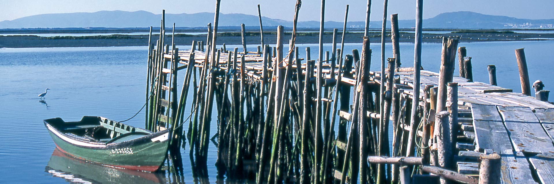 Ancoradouro rustico em madeira com barco amarrado, uma pequena garça na água e montanhas ao fundo.