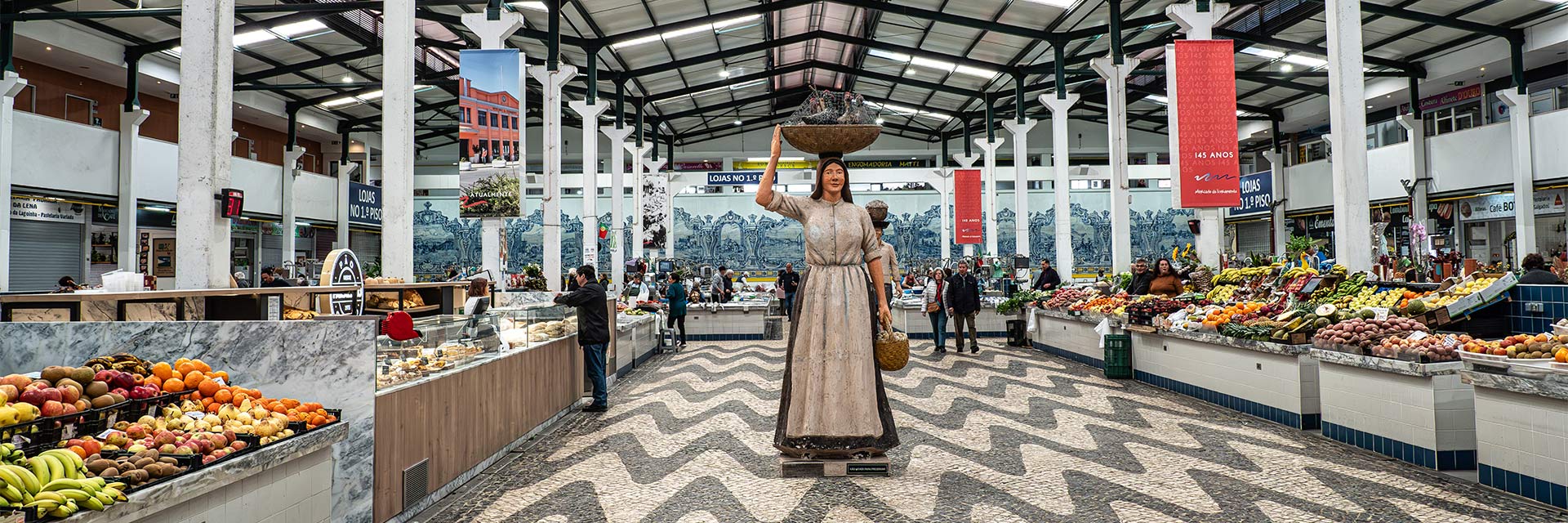 Imagem a cores do interior do Mercado do Livramento, com figura decorativa ao centro e bancas com venda de produtos alimentares.