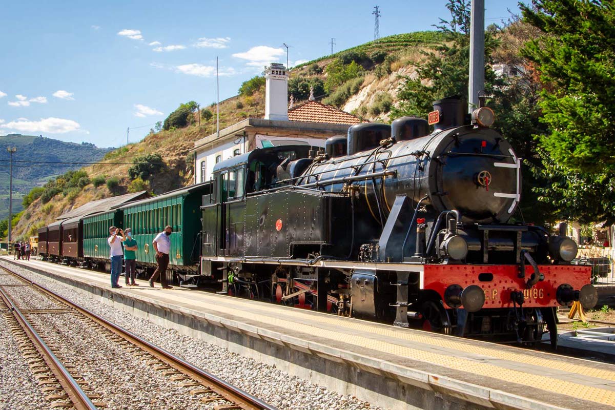 Douro Historical Train, formed by CP 0186 steam locomotive with black and red livery, and five wooden carriages with green and brown livery, stopped at the station.