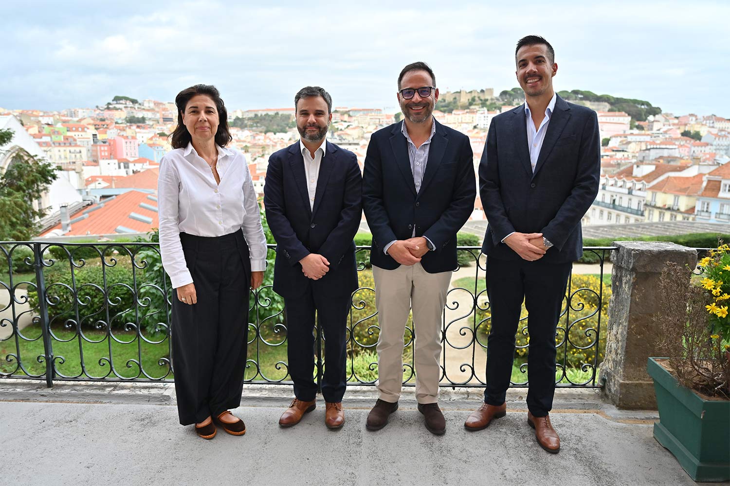 Quatro pessoas lado a lado, com casas e o castelo de São Jorge, em Lisboa, ao fundo.