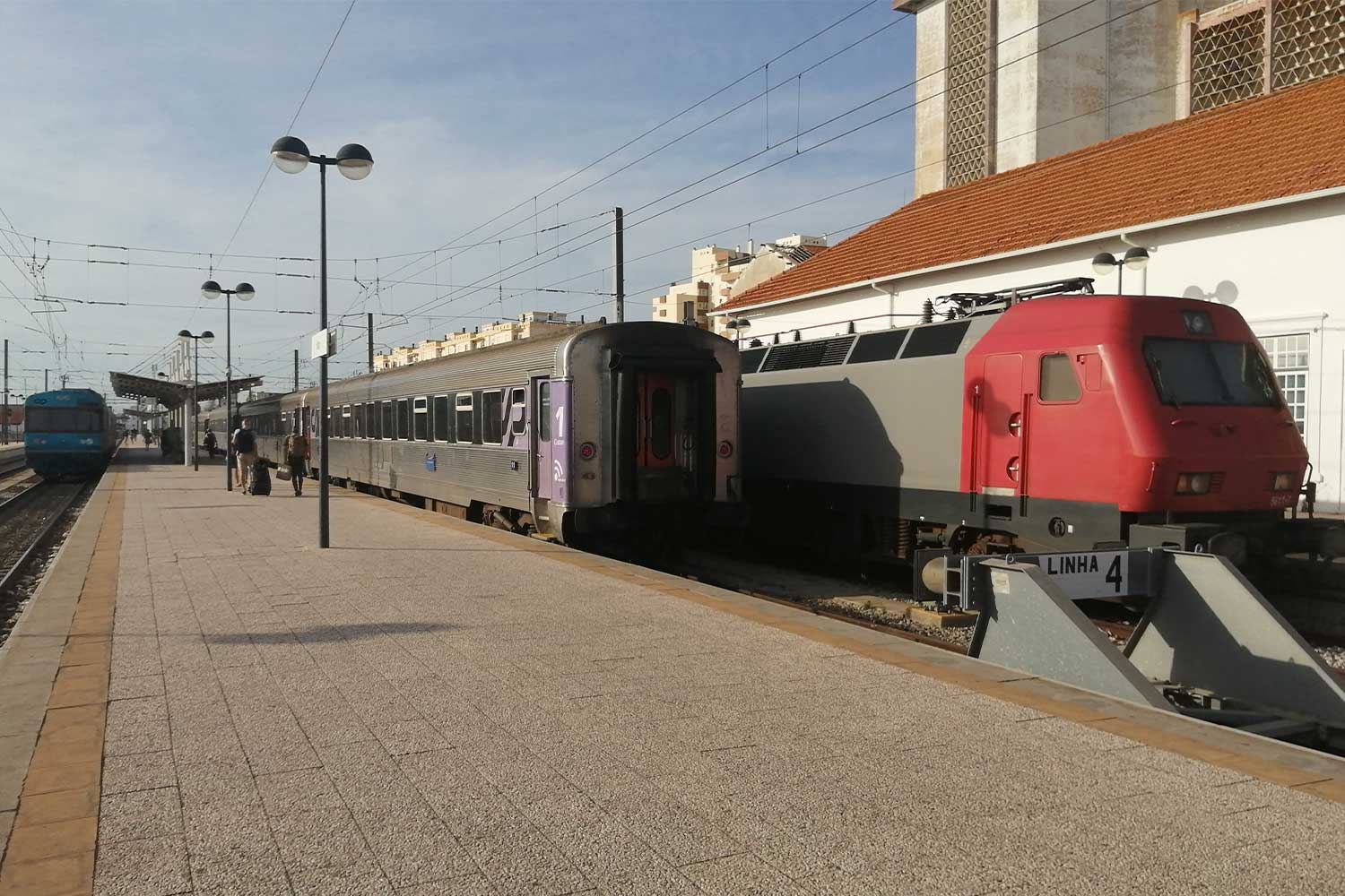 Imagem a cores da Estação de Faro com comboio regional, formado por automotora diesel de cor azul, e comboio Intercidades, formado por locomotiva elétrica da série CP 5600, cores cinzento e vermelho, e carruagens Sorefame, cor cinzento.