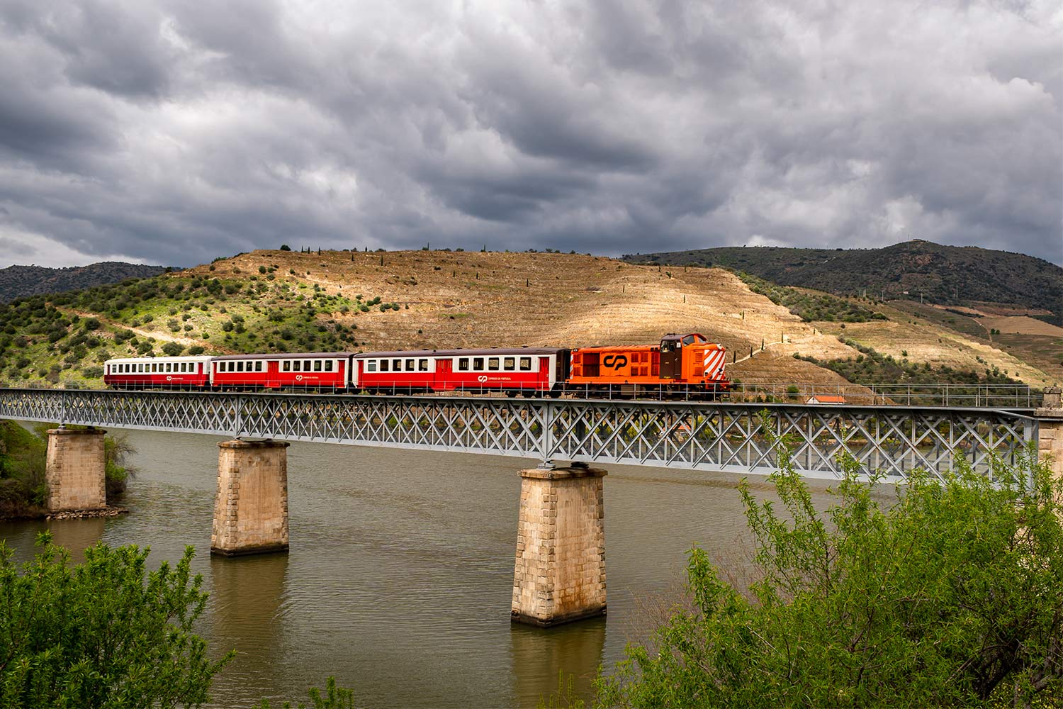 Comboio Miradouro, com locomotiva diesel da série CP 1400, cor laranja e castanho, e carruagens Schindler, cor vermelho e branco, atravessa a ponte ferroviária.
