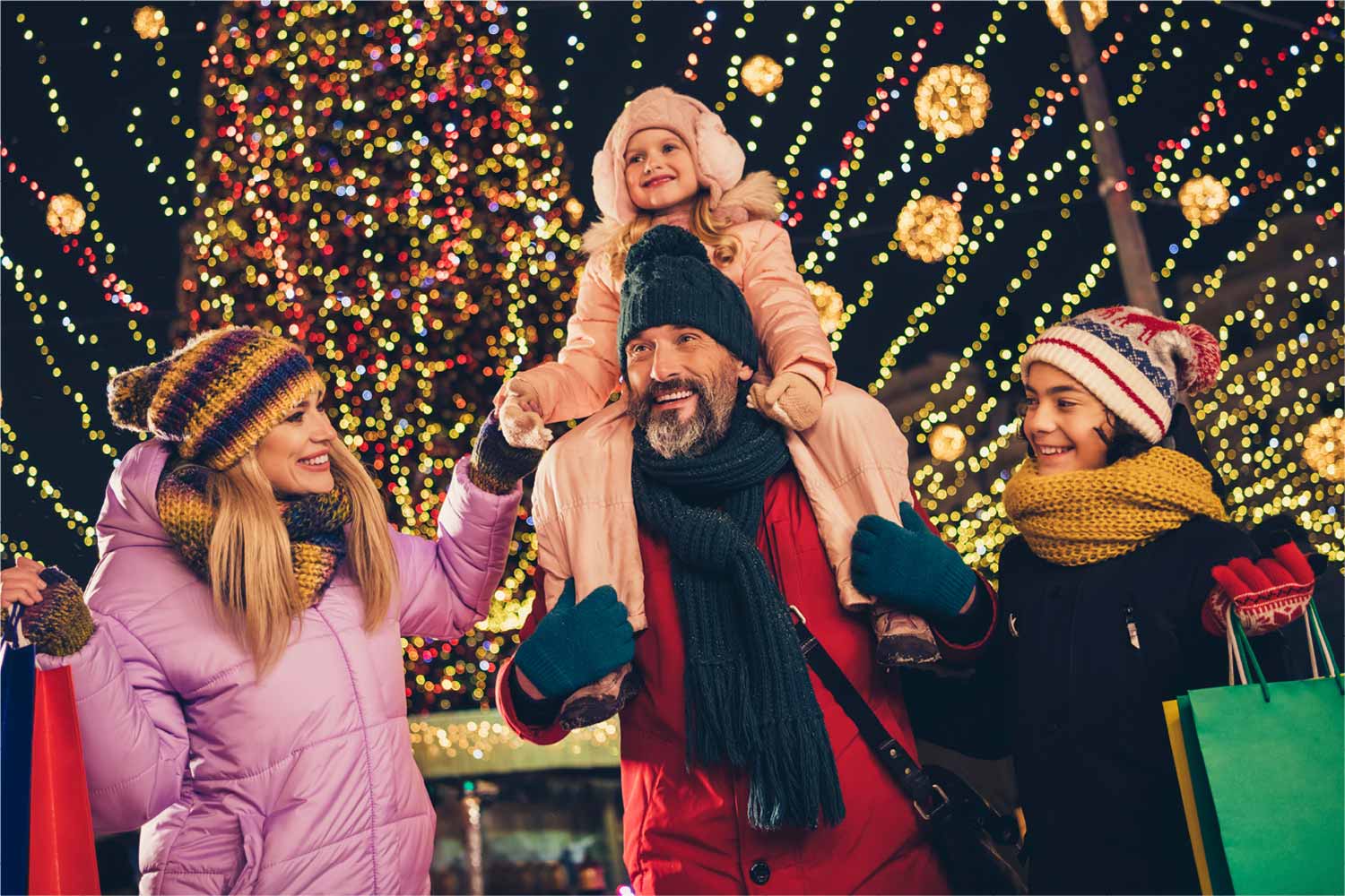 A family of four, with shopping bags, having fun in a space decorated with Christmas lights.