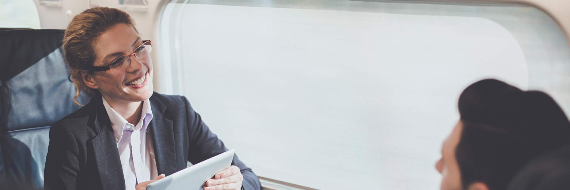 Mulher executiva com tablete e homem ao telemóvel, frente a frente numa mesa de reuniões, junto à janela, no Alfa Pendular.