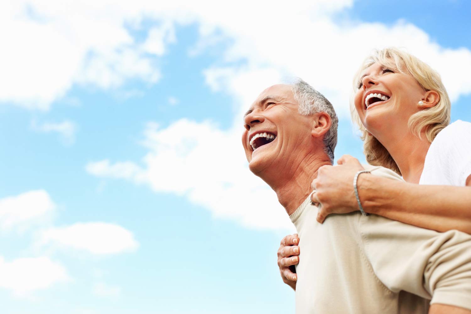 Casal senior abraçado, com céu azul ao fundo.