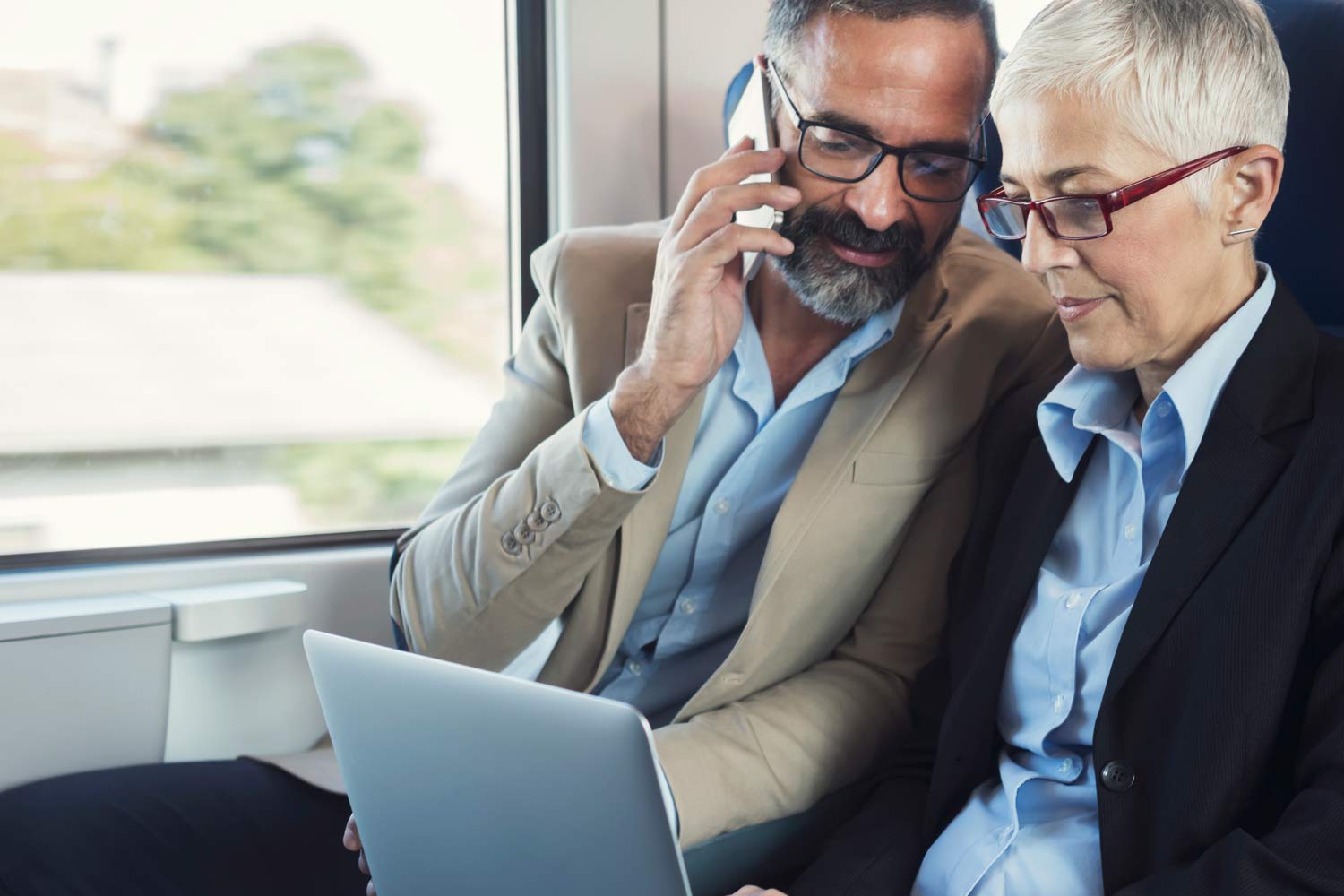  Man on the phone and woman on the laptop, sitting side by side on the train.
      