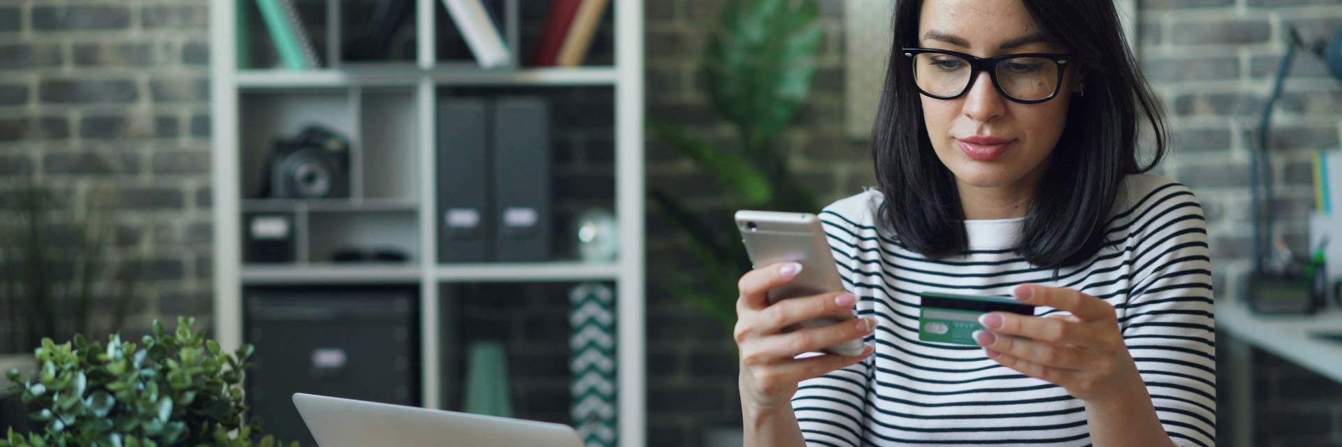 Cliente sentada, com portátil na secretária e de telemóvel e cartão bancário nas mãos.