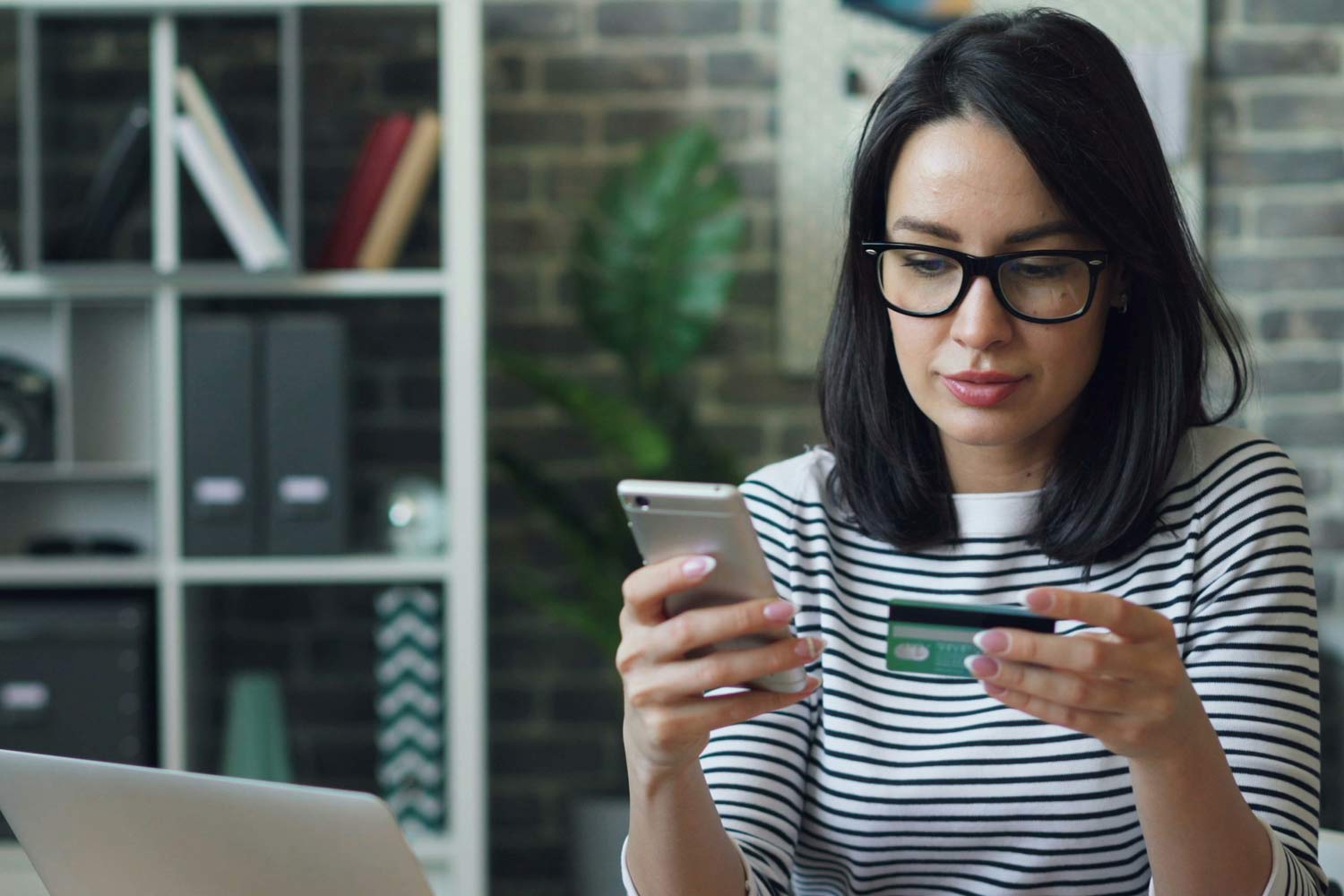 Cliente sentada, com portátil na secretária e de telemóvel e cartão bancário nas mãos.