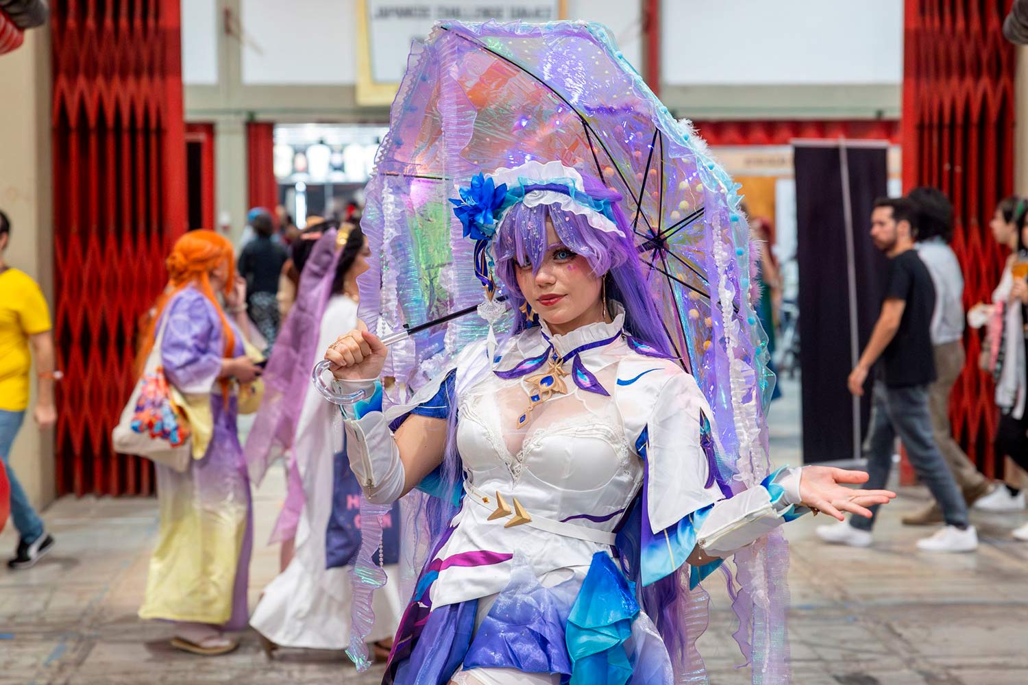 Imagem de rapariga caraterizada e com roupas em tons de branco e roxo com pessoas a passarem por detrás. 