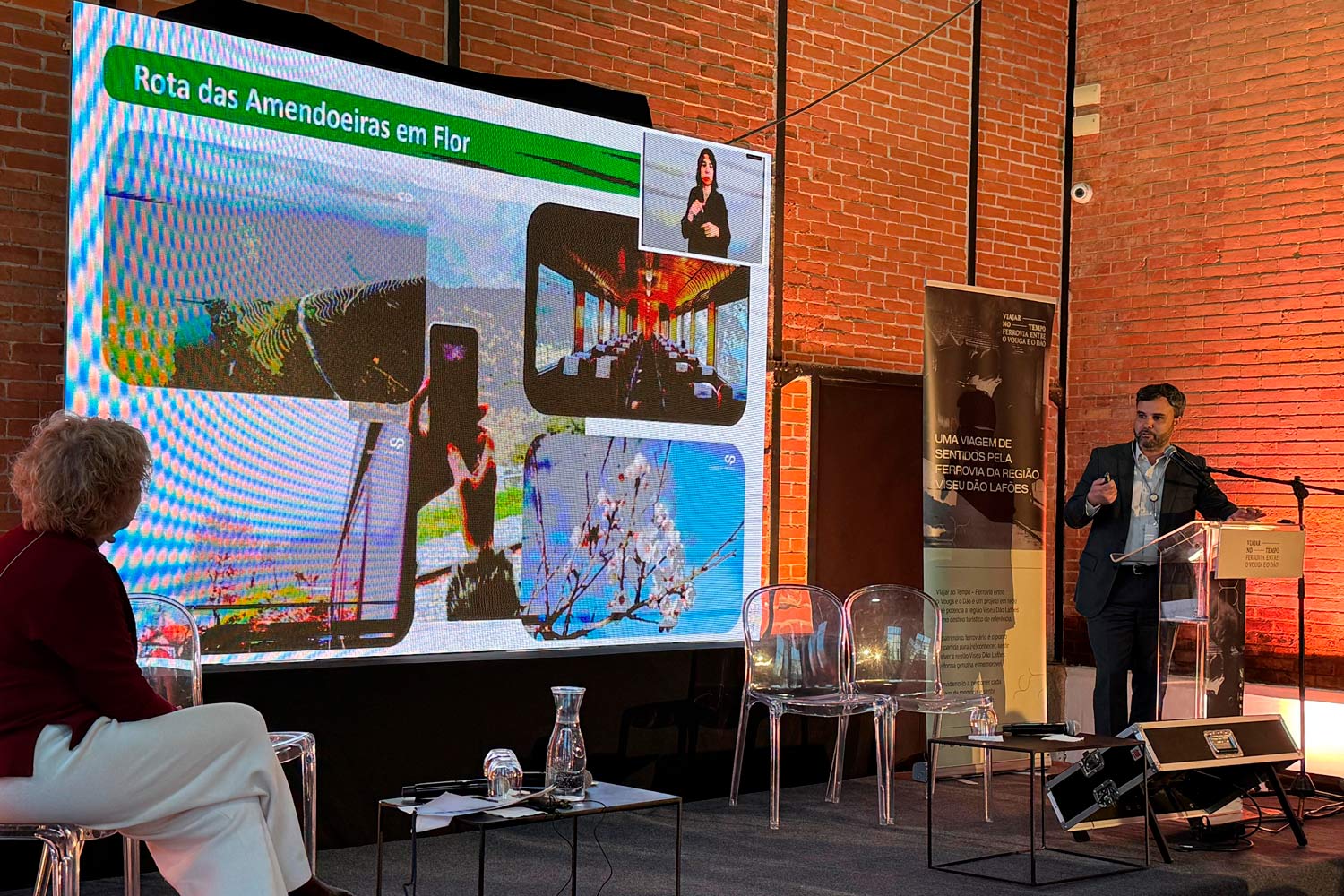 Palco da conferência com o Presidente da CP a apresentar a Rota das Amendoeiras no palco onde também está uma senhora sentada.
